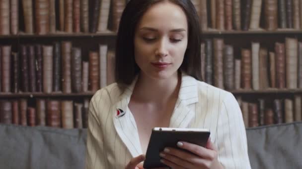 A menina está sentada na biblioteca e lendo um e-book — Vídeo de Stock