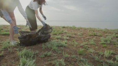 İki kadın siyah bir çöp torbasını kapatıyor. Plastik kirliliği ve çevresel sorun kavramı. Plastikten doğanın gönüllü temizliği.