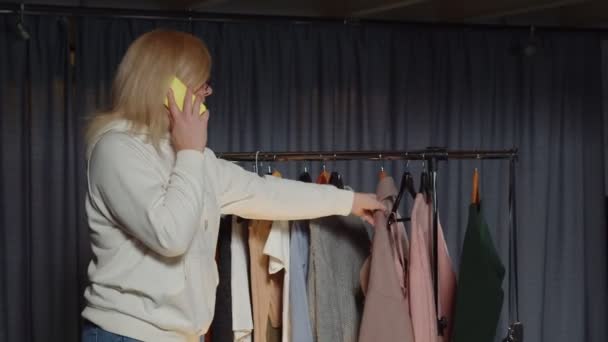 Νεαρή γυναίκα πωλητής μιλάει στο τηλέφωνο και εργάζεται σε κατάστημα μεταχειρισμένων ρούχων — Αρχείο Βίντεο
