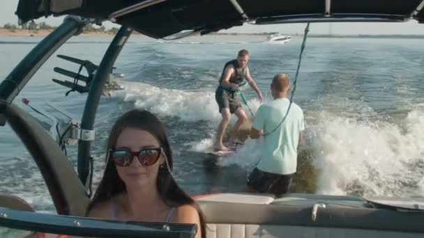 Une femme conduit un bateau à moteur, et en arrière-plan un gars monte un wakeboard, et l'homme l'aide — Video