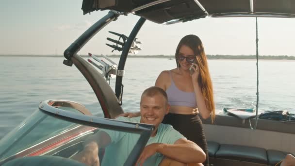 Een man zit in een motorboot, en een vrouw staat naast hem en praat aan de telefoon — Stockvideo