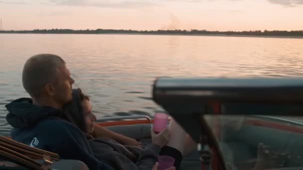 Um cara e uma menina abraçar bonito ao pôr do sol sentado em um barco — Vídeo de Stock