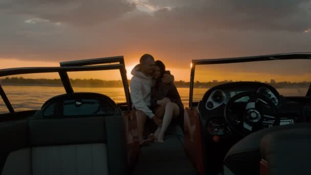 Um tipo e uma rapariga sentados num barco a motor a beijar-se e a ver o pôr-do-sol. Ambiente romântico. — Vídeo de Stock