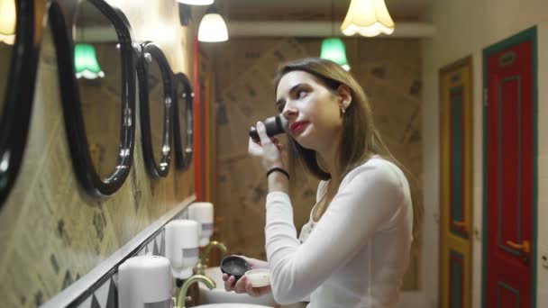 Vista lateral de uma mulher usando pó com uma escova de maquiagem na frente de um espelho, uma mulher feliz usando cosméticos para a beleza — Vídeo de Stock