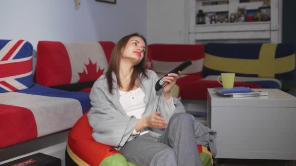 Een jonge vrouw zit 's avonds op een poef in de kamer. Maakt gebruik van de afstandsbediening van de TV, flippen via de kanalen, vindt geen interessante programma 's — Stockvideo