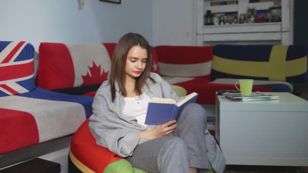 Een jonge vrouw zit thuis op een poef en leest een boek. Een vrouw rust binnen op een comfortabele poef in haar vrije tijd. Heb een leuke vrije tijd in het weekend — Stockvideo