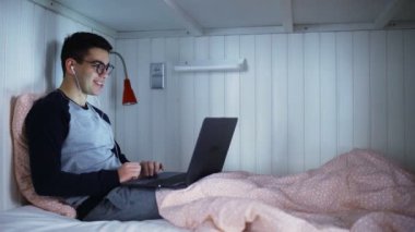 Yandan bakıldığında genç bir adam yatakta yatar ve dizüstü bilgisayarla çalışır, klavyeye yazar, geceleri çalışır, gece evden uzaktan kumandayla çalışır..