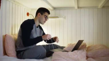 Yatakta genç bir adam oturuyor ve bir dizüstü bilgisayarla çalışıyor, kulaklıkları takıyor ve klavyeye yazıyor, geceleri çalışıyor, uzaktan kumandayla evden çalışıyor..