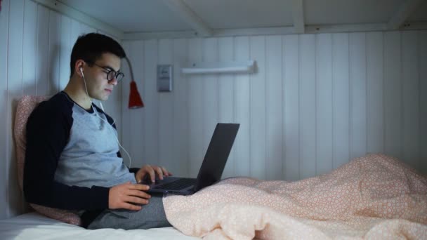 Vista lateral un hombre joven está acostado en una cama y trabajando en un ordenador portátil, escribiendo en un teclado, trabajando por la noche, trabajo remoto desde casa por la noche. — Vídeos de Stock