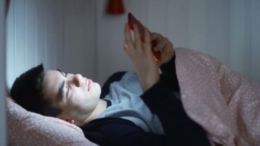 Yatakta kulaklık takmış ve gece biriyle mesajlaşan genç bir adamın yakın çekimi. Gece yazışmaları