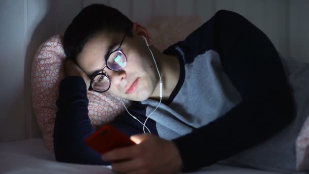 Close-up de um jovem deitado na cama com fones de ouvido e mensagens de texto com alguém à noite. Correspondência nocturna — Vídeo de Stock