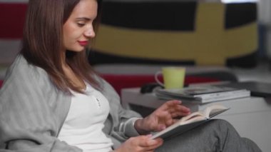 Genç bir kadın evinde bir sedirin üzerinde oturuyor ve kitap okuyor. Bir kadın boş zamanlarında rahat bir sedirin üzerinde dinleniyor. Hafta sonları iyi vakit geçir.