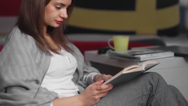 Молодая женщина сидит дома на оттоманке и читает книгу. Женщина отдыхает в помещении на удобной оттоманке в свободное время. Хорошо провести свободное время в выходные — стоковое видео
