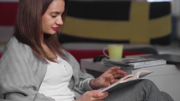 Una joven está sentada en casa en una otomana y leyendo un libro. Una mujer descansa en un cómodo otomano en su tiempo libre. Tener un buen tiempo libre en el fin de semana — Vídeos de Stock