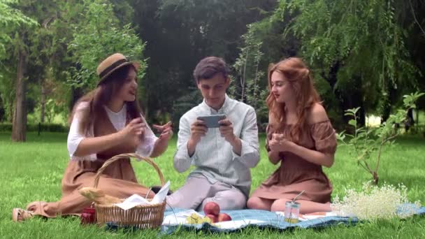 El chico juega el juego en el dispositivo, y las chicas a su lado lo apoyan en la naturaleza en el verano — Vídeos de Stock