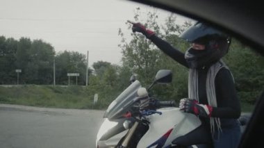 Siyah kasklı genç bir kadın motosiklette oturur ve araba şoförüyle konuşur.
