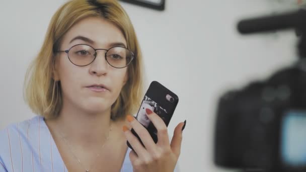 Detailní záběr mladé ženy s brýlemi, která sedí s telefonem v ruce, usmívá se a mluví do kamery — Stock video