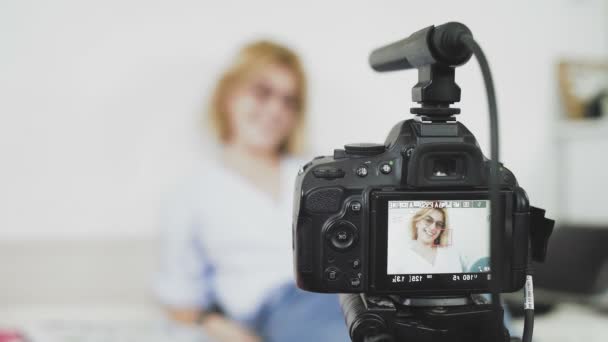 Een jonge vrouw met een bril zet de camera aan, lacht en praat met de camera. Groeten aan uw kijkers — Stockvideo