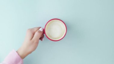Pastel mavi arka planda bir fincan kahvenin üst görüntüsü. Bir kadının eli, bir fincan kahveyi pastel mavi arka plandan kaldırır.