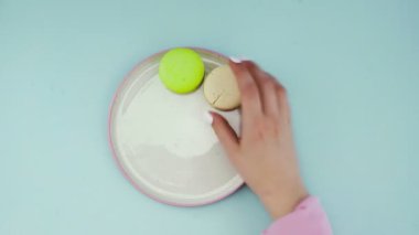 Pastel mavi arka planda çok renkli Fransız makarnalı kurabiyelerin üst görüntüsü. Bir kadının eli tabağa kurabiye koyar.