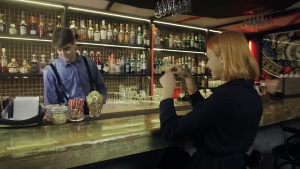 Дівчина сидить у барі з коктейлем у руках і спілкується з барменом — стокове відео