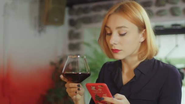 Uma menina com um copo de vinho em um bar está sentada em uma mesa de bar, bebendo vinho e escrevendo mensagens — Vídeo de Stock
