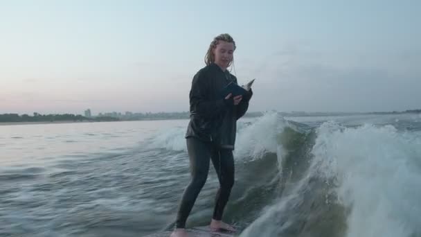 Samica surferki wskakuje na wakeboard i czyta książkę. Doświadczony wakeboarder spryskuje kamerą krople wody. — Wideo stockowe