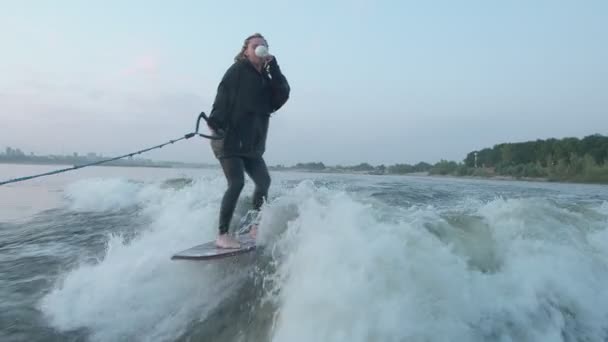 Samica surferki wskakuje na wakeboard i pije kawę lub herbatę z filiżanki. Doświadczony wakeboarder spryskuje kamerą krople wody. — Wideo stockowe