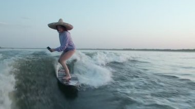 Hasır şapkalı bir sörfçü elinde telefonla bir sörf tahtasına atlıyor. Deneyimli bir wakeboardcu kameraya su püskürtür..