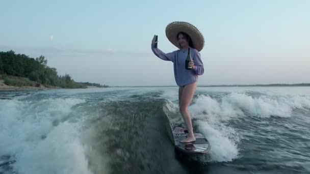 Μια γυναίκα σέρφερ με ψάθινο καπέλο πηδάει σε μια σανίδα αφύπνισης με σαμπάνια στα χέρια της, βγάζοντας μια σέλφι στο κινητό της. Ένας έμπειρος wakeboarder ψεκάζει νερό σταγόνες στην κάμερα. — Αρχείο Βίντεο
