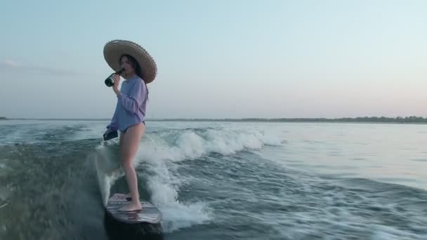 Ένας σέρφερ με ψάθινο καπέλο πηδάει σε μια σανίδα με σαμπάνια και ένα τηλέφωνο στα χέρια της. Ένας έμπειρος wakeboarder ψεκάζει νερό σταγόνες στην κάμερα. — Αρχείο Βίντεο