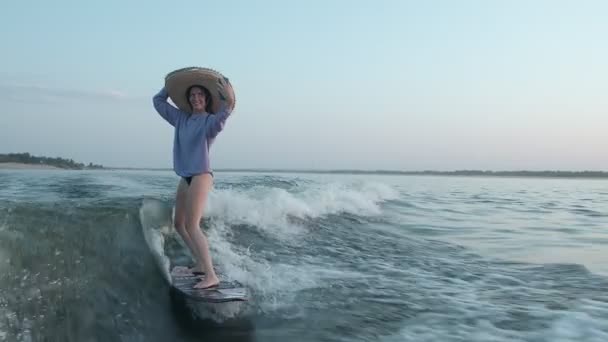 Ένας σέρφερ με ψάθινο καπέλο πηδάει σε μια σανίδα και βγάζει μια σέλφι στο κινητό της. Ένας έμπειρος wakeboarder ψεκάζει νερό σταγόνες στην κάμερα. — Αρχείο Βίντεο