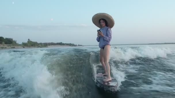 Una surfista con un sombrero de paja salta sobre un wakeboard con un teléfono en sus manos. Un experimentado wakeboarder rocía agua en la cámara. — Vídeos de Stock