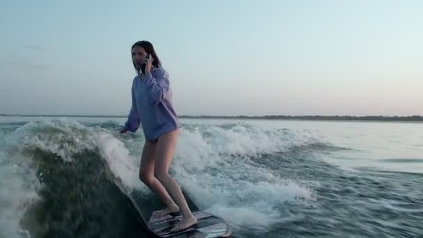 Surfer skacze na desce z telefonem w rękach. Doświadczony wakeboarder spryskuje kamerą krople wody. — Wideo stockowe