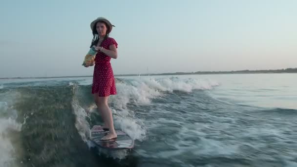 Une surfeuse en robe rouge et chapeau saute sur un wakeboard et tient un paquet de chips dans ses mains et mange. Un wakeboarder expérimenté pulvérise de l'eau dans la caméra. — Video