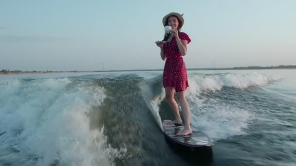 Um surfista em um vestido vermelho e chapéu pula em um wakeboard e segura uma xícara de chá ou café em suas mãos. Um wakeboarder experiente pulveriza água cai na câmera. — Vídeo de Stock
