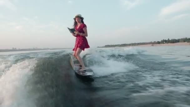 Surfařka v červených šatech a klobouku skočí na wakeboard a drží knihu v ruce. Zkušený wakeboarder rozstřikuje do kamery kapky vody. — Stock video