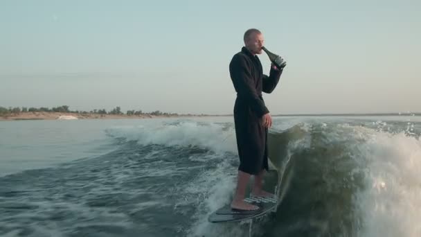 Surfař skákající na wakeboardu v froté županu s lahví šampaňského. Zkušený wakeboarder rozstřikuje do kamery kapky vody. — Stock video