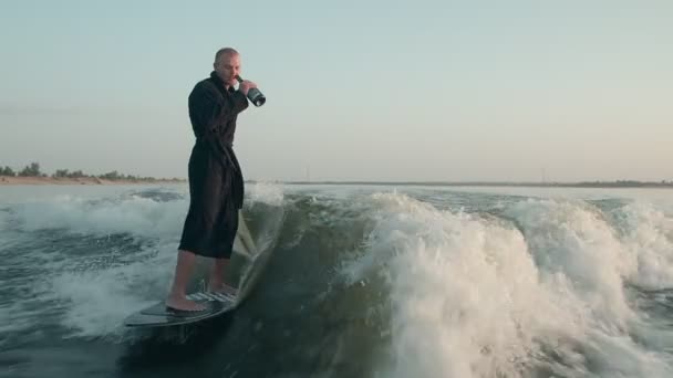 Surfař skákající na wakeboardu v froté županu s lahví šampaňského. Zkušený wakeboarder rozstřikuje do kamery kapky vody. — Stock video