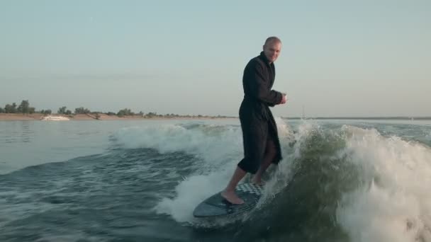Surfař skákající na wakeboardu v froté županu s šálkem čaje nebo kávy v ruce. Zkušený wakeboarder rozstřikuje do kamery kapky vody. — Stock video
