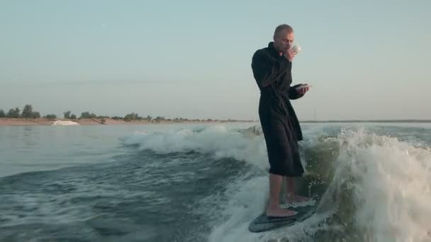 Surfař skákající na wakeboardu v froté županu s šálkem čaje nebo kávy v ruce. Zkušený wakeboarder rozstřikuje do kamery kapky vody. — Stock video