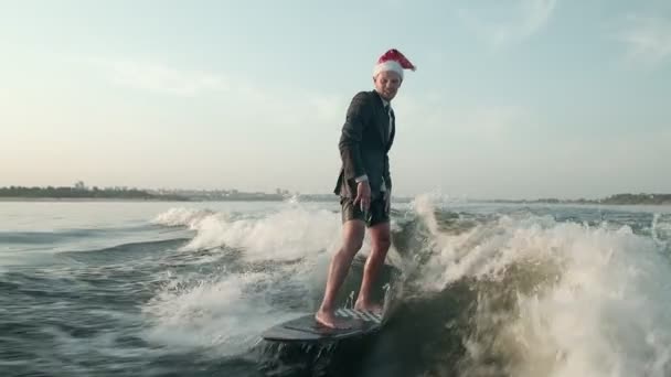 サーファーはスーツを着たウェイクボードに飛び乗り、頭にはクリスマスの赤い帽子をかぶっている。経験豊富なウェイクボーダーがカメラに水を噴霧. — ストック動画