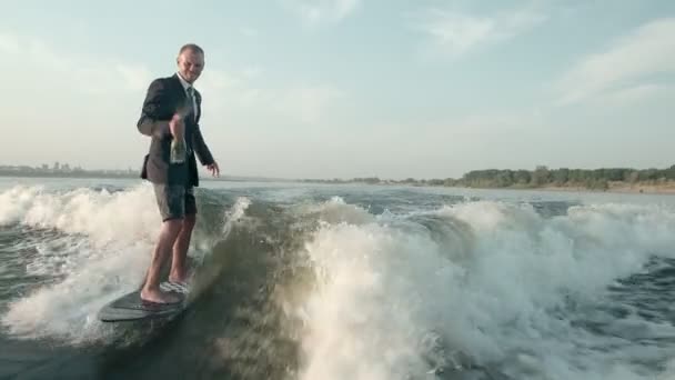 Ένας σέρφερ που πηδάει πάνω σε μια σανίδα με κοστούμι και ένα μπουκάλι σαμπάνια. Ένας έμπειρος wakeboarder ψεκάζει νερό σταγόνες στην κάμερα. — Αρχείο Βίντεο