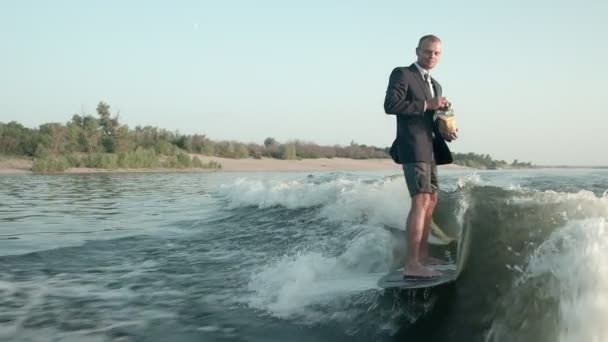 Bir sörfçü takım elbiseyle bir sörf tahtasına atlıyor ve elinde bir paket cips tutuyor. Sefer cips yer ve wakeboard kullanır. Deneyimli bir wakeboardcu kameraya su püskürtür.. — Stok video
