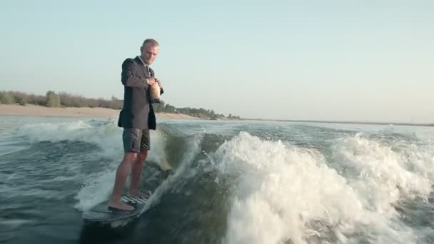 Surfer skaczący na desce w garniturze i trzymający w rękach paczkę żetonów. Sefer je chipsy i jeździ na wakeboardzie. Doświadczony wakeboarder spryskuje kamerą krople wody. — Wideo stockowe