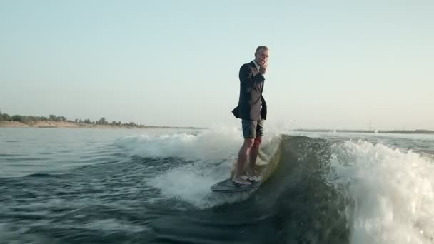 Um surfista a saltar num wakeboard de fato com um pão nas mãos. O surfista come um pão e monta um wakeboard. Um wakeboarder experiente pulveriza água cai na câmera. — Vídeo de Stock