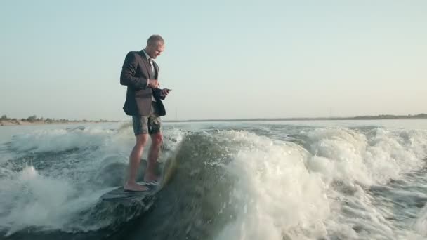 Un surfista saltando en un wakeboard en un traje con un teléfono en sus manos. Un experimentado wakeboarder rocía agua en la cámara. — Vídeos de Stock