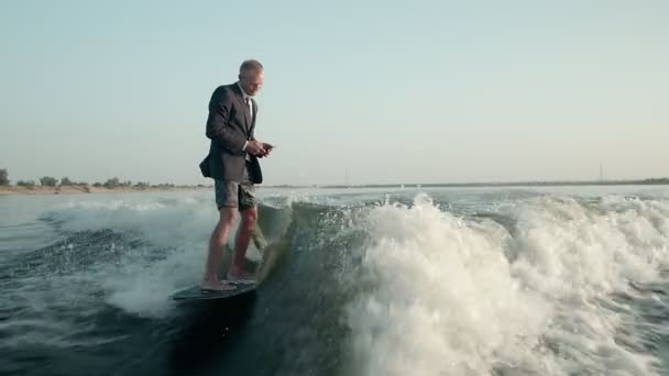 サーファーが携帯電話を手にスーツを着てウェイクボードにジャンプします。経験豊富なウェイクボーダーがカメラに水を噴霧. — ストック動画