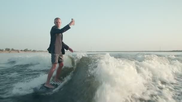 Surfař skákající na wakeboardu v obleku s telefonem v ruce si sám sebe fotí. Surfař si vezme selfie a jede na wakeboardu. — Stock video