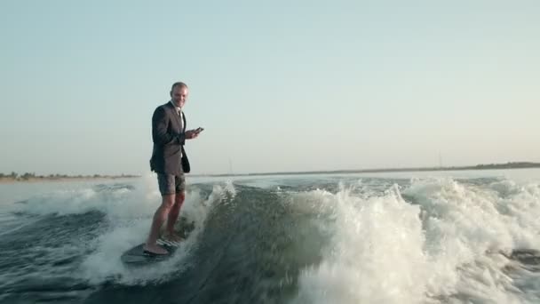 Surfař skákající na wakeboardu v obleku s telefonem v ruce. Zkušený wakeboarder rozstřikuje do kamery kapky vody. — Stock video